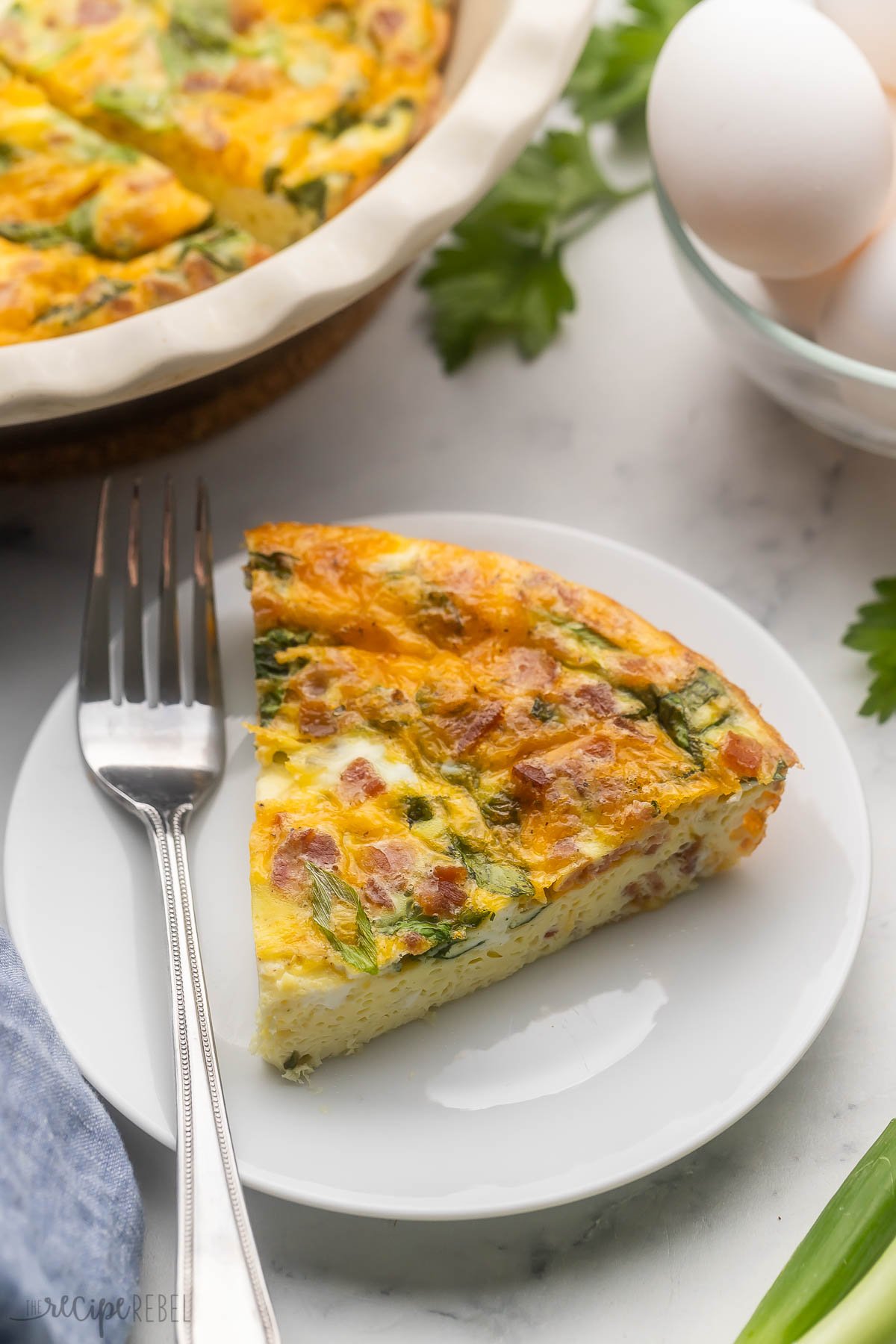 Close up view of a slice of Crustless Quiche on a white plate with a fork next to it.